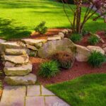 Well-designed garden showcasing landscape design with stone steps, lush green lawn, assorted perennials, and a young tree.